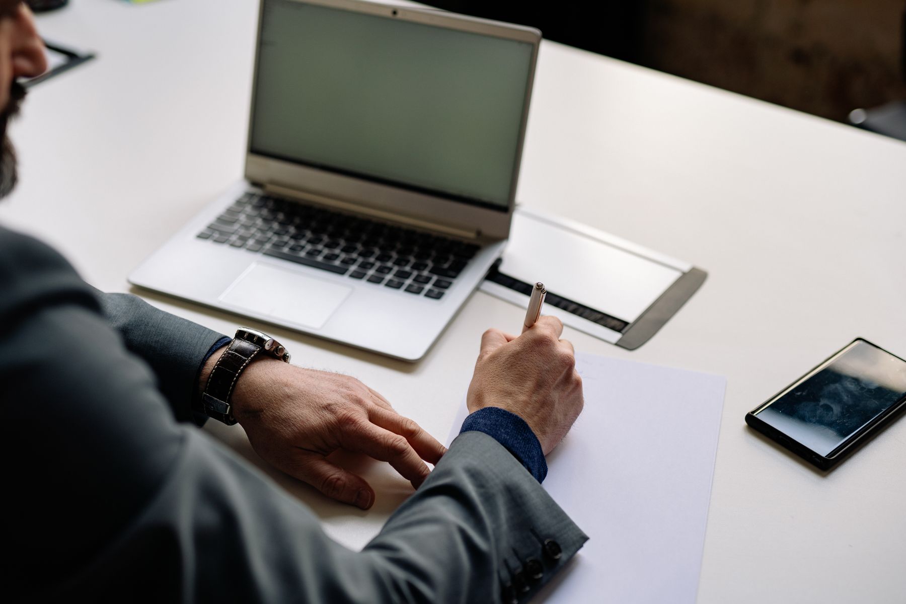 A busy professional making notes at a desk beside a laptop and phone