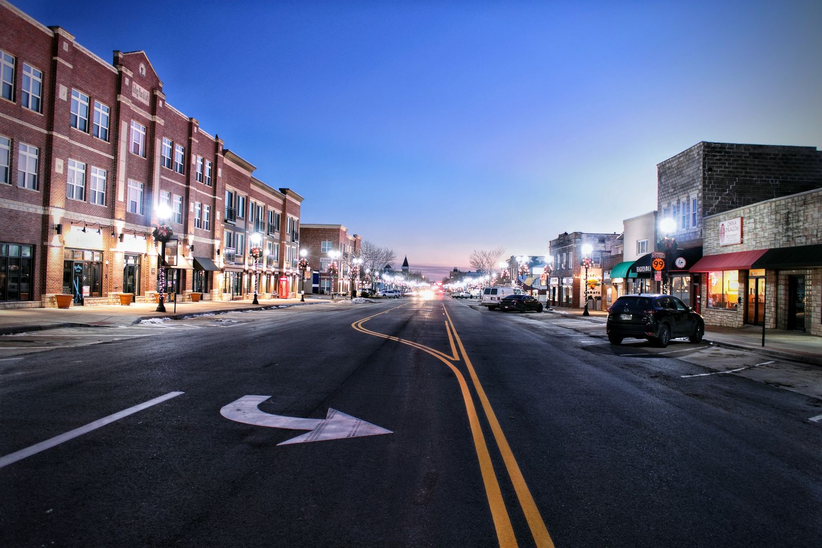 A small-town American main street at twilight, storefronts lit and the road empty