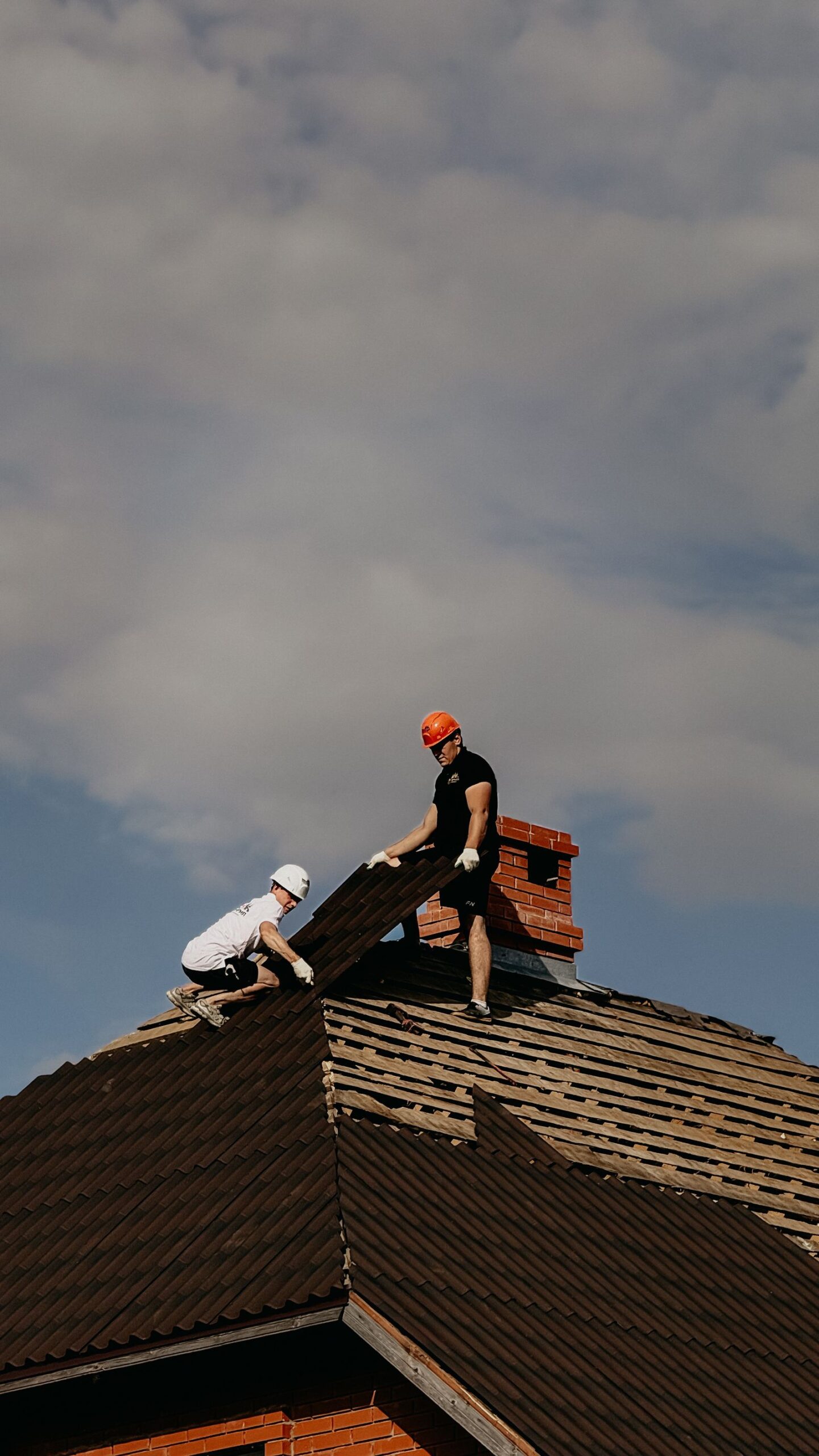 Two roofers in safety helmets working on a house roof under a cloudy sky