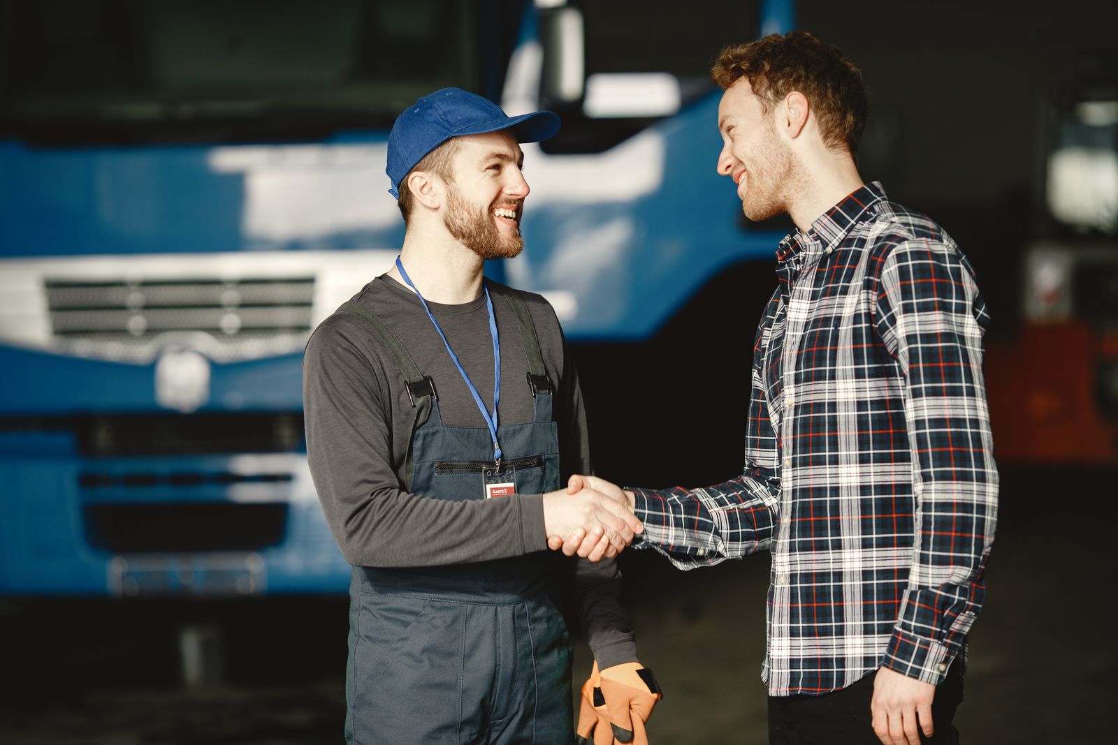 A mechanic in overalls shaking hands with a customer in front of a truck in a garage
